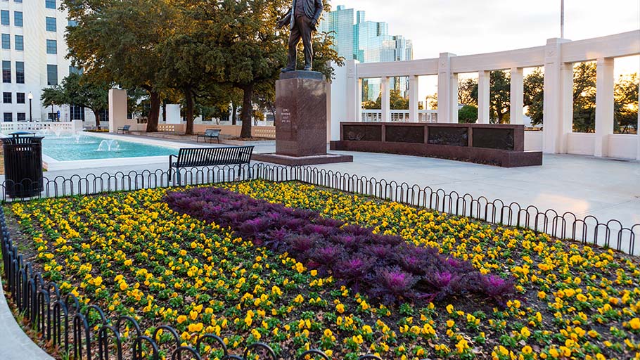 An image of a public plaza with a large fountain, garden, and a tall statue of a man