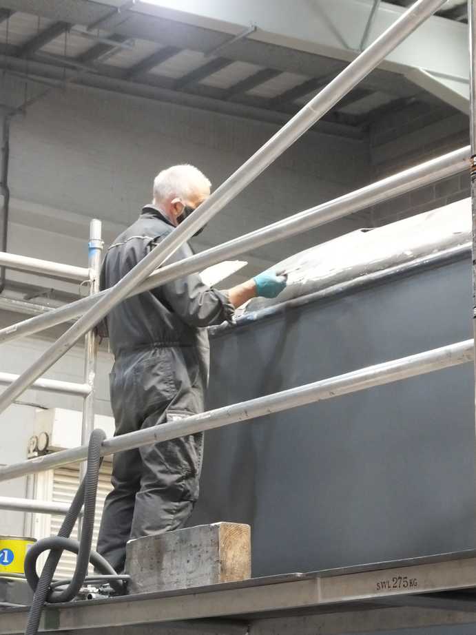 A man in grey overalls stands on a scaffold, sanding a locomotive tender